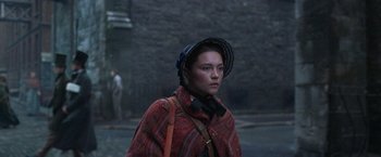 Movie still from “The Wonder” (2022), directed by Sebastián Lelio – A woman wearing a hat is standing on the street; Medium shot, Low angle