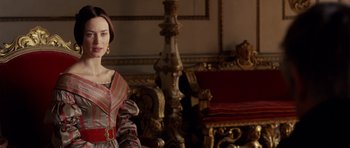Movie still from “The Young Victoria” (2009), directed by Jean-Marc Vallée – A woman in a red and white dress sitting in front of a red couch; Medium shot, Over the shoulder angle