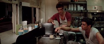 Movie still from “Scarface” (1983), directed by Brian De Palma – A man pouring a drink into a cup; Medium shot, Over the shoulder angle