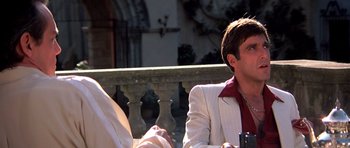Movie still from “Scarface” (1983), directed by Brian De Palma – A man in a white suit sitting next to another man in a white suit; Close Up shot, Low angle