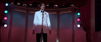 Movie still from “Scarface” (1983), directed by Brian De Palma – A man in a white suit is speaking on a microphone; Medium shot, Low angle