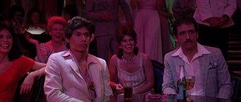 Movie still from “Scarface” (1983), directed by Brian De Palma – A man and a woman sitting at a table in front of other people; Medium shot, Over the shoulder angle