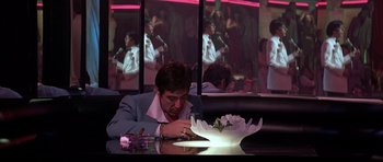 Movie still from “Scarface” (1983), directed by Brian De Palma – A man sitting at a table with a bowl of flowers in front of him; Wide shot, Over the shoulder angle