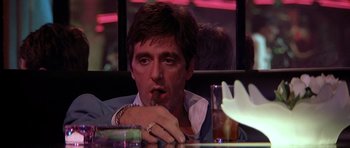 Movie still from “Scarface” (1983), directed by Brian De Palma – A person sitting at a table smoking a cigarette; Close Up shot, Low angle
