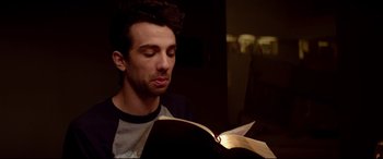 Movie still from “This Is the End” (2013), directed by Seth Rogen – A young man reading a book in a dark room; Close Up shot, Over the shoulder angle