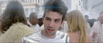 Movie still from “This Is the End” (2013), directed by Seth Rogen – A young man in a silver shirt is standing in front of a group of people; Close Up shot, Over the shoulder angle