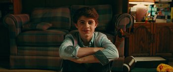 Movie still from “To All the Boys I've Loved Before” (2018), directed by Susan Johnson – A young man sitting on top of a couch with his arms crossed; Medium shot, Low angle
