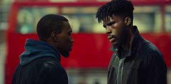 Movie still from “Top Boy” (2011), created by Ronan Bennett – Two men standing next to each other in front of a red bus; Close Up shot, Over the shoulder angle
