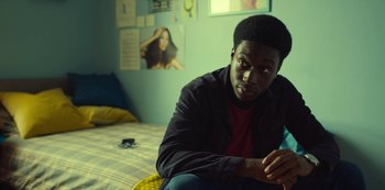 Movie still from “Top Boy” (2011), created by Ronan Bennett – A man sitting on a bed in front of a wall; Medium shot, Over the shoulder angle