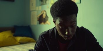 Movie still from “Top Boy” (2011), created by Ronan Bennett – A man sitting in front of a wall with pictures on the wall behind him; Close Up shot, Over the shoulder angle