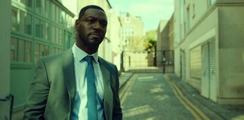Movie still from “Top Boy” (2011), created by Ronan Bennett – A man wearing a suit and tie standing on a street; Close Up shot, Over the shoulder angle