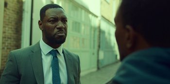 Movie still from “Top Boy” (2011), created by Ronan Bennett – A man wearing a suit and tie standing next to another man; Close Up shot, Over the shoulder angle