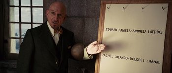 Movie still from “Shutter Island” (2010), directed by Martin Scorsese – A man in a suit and bow tie pointing to a board with names on it; Medium shot, Overhead angle