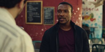 Movie still from “Top Boy” (2011), created by Ronan Bennett – A man in a black jacket is standing in front of a chalkboard; Close Up shot, Over the shoulder angle