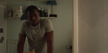 Movie still from “Top Boy” (2011), created by Ronan Bennett – A man standing next to a glass of orange juice in a living room; Medium shot, Over the shoulder angle