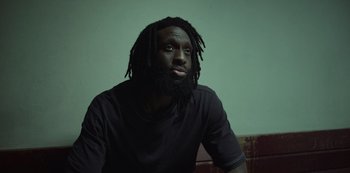 Movie still from “Top Boy” (2011), created by Ronan Bennett – A man with dreads sitting in front of a green wall; Close Up shot, Low angle