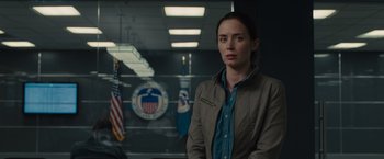 Movie still from “Sicario” (2015), directed by Denis Villeneuve – A woman standing in front of an american flag in an office; Medium shot, Over the shoulder angle