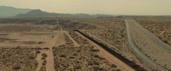 Movie still from “Sicario” (2015), directed by Denis Villeneuve – An aerial view of a dirt road with a fence on the side of it; Extreme Wide shot, High angle
