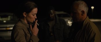 Movie still from “Sicario” (2015), directed by Denis Villeneuve – A woman smoking a cigarette while two other people watch; Medium shot, Over the shoulder angle