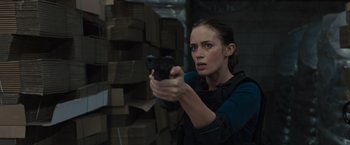 Movie still from “Sicario” (2015), directed by Denis Villeneuve – A woman holding a gun while standing next to a wall; Medium shot, Over the shoulder angle