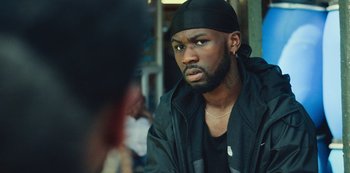 Movie still from “Top Boy” (2011), created by Ronan Bennett – A man in a black jacket and a black hat looking at the camera; Close Up shot, Over the shoulder angle