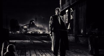 Movie still from “Sin City” (2005), directed by Frank Miller – A black and white photo of a man in a trench coat; Wide shot, Low angle