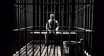 Movie still from “Sin City” (2005), directed by Frank Miller – A man sitting in a jail cell with his hands on his hips; Wide shot, High angle