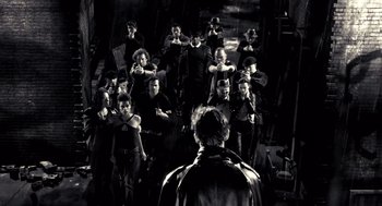 Movie still from “Sin City” (2005), directed by Frank Miller – A group of people standing around a man in a black jacket; Wide shot, High angle