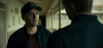 Movie still from “Triple Frontier” (2019), directed by J.C. Chandor – A man wearing a baseball cap talking to another man in front of a building; Close Up shot, Over the shoulder angle