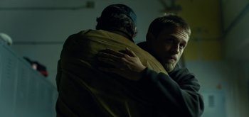 Movie still from “Triple Frontier” (2019), directed by J.C. Chandor – A man is hugging another man in a dark room; Medium shot, Over the shoulder angle