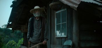 Movie still from “Triple Frontier” (2019), directed by J.C. Chandor – An old man standing in front of a wooden house; Medium shot, Low angle
