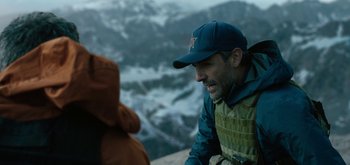 Movie still from “Triple Frontier” (2019), directed by J.C. Chandor – A man wearing a hat and a jacket looking at a teddy bear on top of a mountain; Close Up shot, Over the shoulder angle