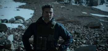 Movie still from “Triple Frontier” (2019), directed by J.C. Chandor – A man wearing a body armor standing on a rocky ground; Medium shot, Over the shoulder angle