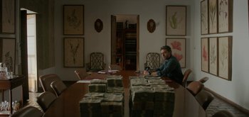 Movie still from “Triple Frontier” (2019), directed by J.C. Chandor – A man sitting at a wooden table in front of stacks of money; Wide shot, Low angle