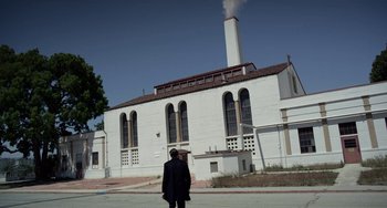 Movie still from “Velvet Buzzsaw” (2019), directed by Dan Gilroy – A man standing in front of a white building with smoke coming out of it; Extreme Wide shot, Low angle