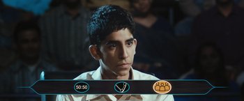 Movie still from “Slumdog Millionaire” (2008), directed by Danny Boyle – A young man is sitting in front of a group of people; Close Up shot, High angle