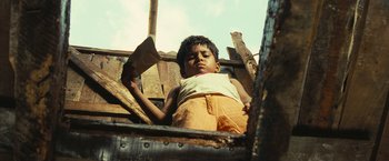 Movie still from “Slumdog Millionaire” (2008), directed by Danny Boyle – A young boy sitting on a wooden platform; Medium shot, Low angle