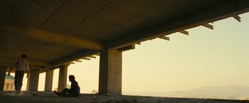 Movie still from “Slumdog Millionaire” (2008), directed by Danny Boyle – A person sitting under a large concrete structure; Extreme Wide shot, Low angle
