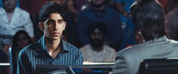 Movie still from “Slumdog Millionaire” (2008), directed by Danny Boyle – A man sitting in front of a group of people; Close Up shot, Over the shoulder angle