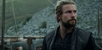Movie still from “Vikings: Valhalla” (2022), created by Jeb Stuart – A man with long hair and a beard is standing in front of some rocks; Close Up shot, Low angle