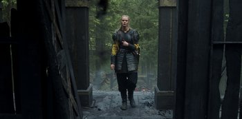 Movie still from “Vikings: Valhalla” (2022), created by Jeb Stuart – A man in black and yellow jacket standing in front of trees; Medium shot, Low angle