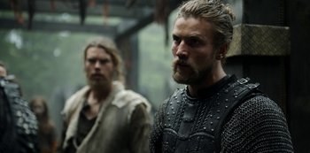 Movie still from “Vikings: Valhalla” (2022), created by Jeb Stuart – A man with long hair and a beard standing next to another man with long hair; Close Up shot, Low angle