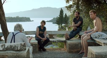 Movie still from “Before Midnight” (2013), directed by Richard Linklater – A group of people sitting next to each other on a stone bench; Medium shot, Over the shoulder angle