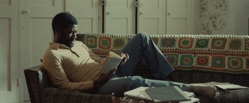 Movie still from “Small Axe” (2020), directed by Steve McQueen – A man sitting on a couch reading a book; Medium shot, Low angle