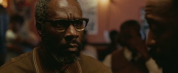 Movie still from “Small Axe” (2020), directed by Steve McQueen – A person wearing glasses and a suit; Close Up shot, Over the shoulder angle