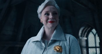 Movie still from “Wednesday” (2022), created by Miles Millar – An older woman wearing a white coat and a gold brooch; Close Up shot, Low angle