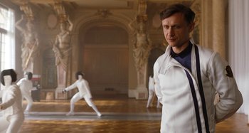 Movie still from “Wednesday” (2022), created by Miles Millar – A man standing in front of a wall with a fencer in the background; Medium shot, Low angle
