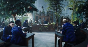Movie still from “Wednesday” (2022), created by Miles Millar – A group of people sitting at tables in a room with plants; Wide shot, High angle