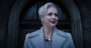Movie still from “Wednesday” (2022), created by Miles Millar – An older woman wearing a gray coat and a gold necklace; Close Up shot, Low angle