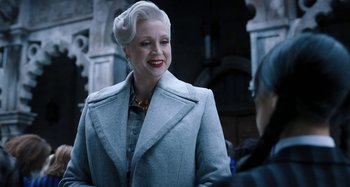 Movie still from “Wednesday” (2022), created by Miles Millar – An older woman in a gray coat and a yellow necklace; Medium shot, Over the shoulder angle